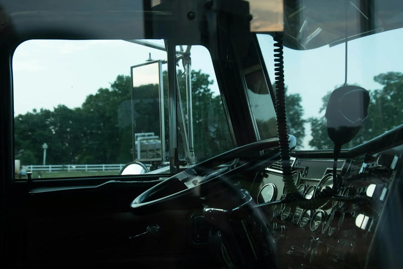 Inside a parked truck cab: the steering wheel, the gauges and a CB microphone hanging from the dash, with trees outside the windshield.